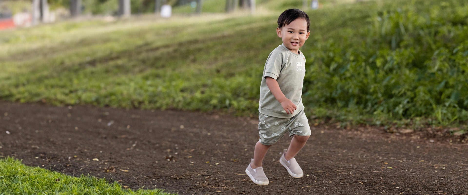 A young boy wearing a green t-shirt and shorts happily running on a dirt path in a park.