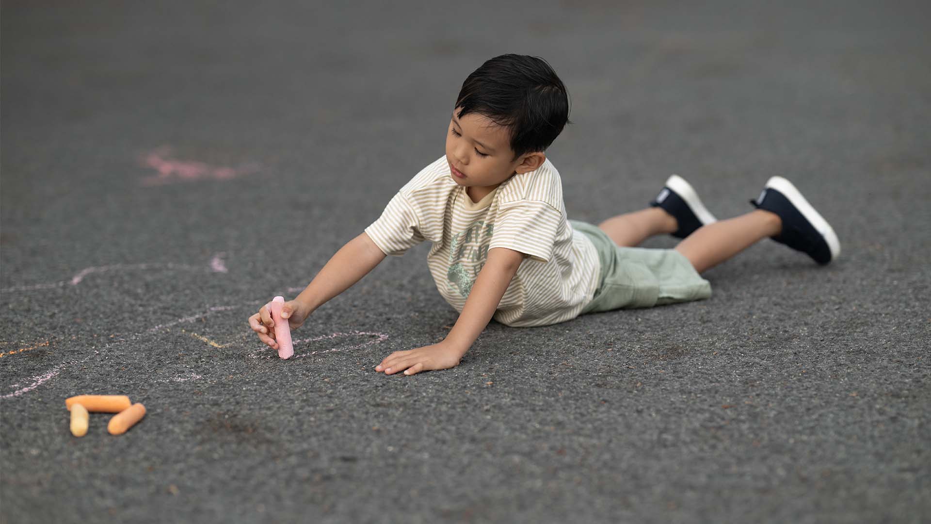  A toddler boy drawing with chalk on pavement, wearing a striped t-shirt and shorts, lying on his stomach.