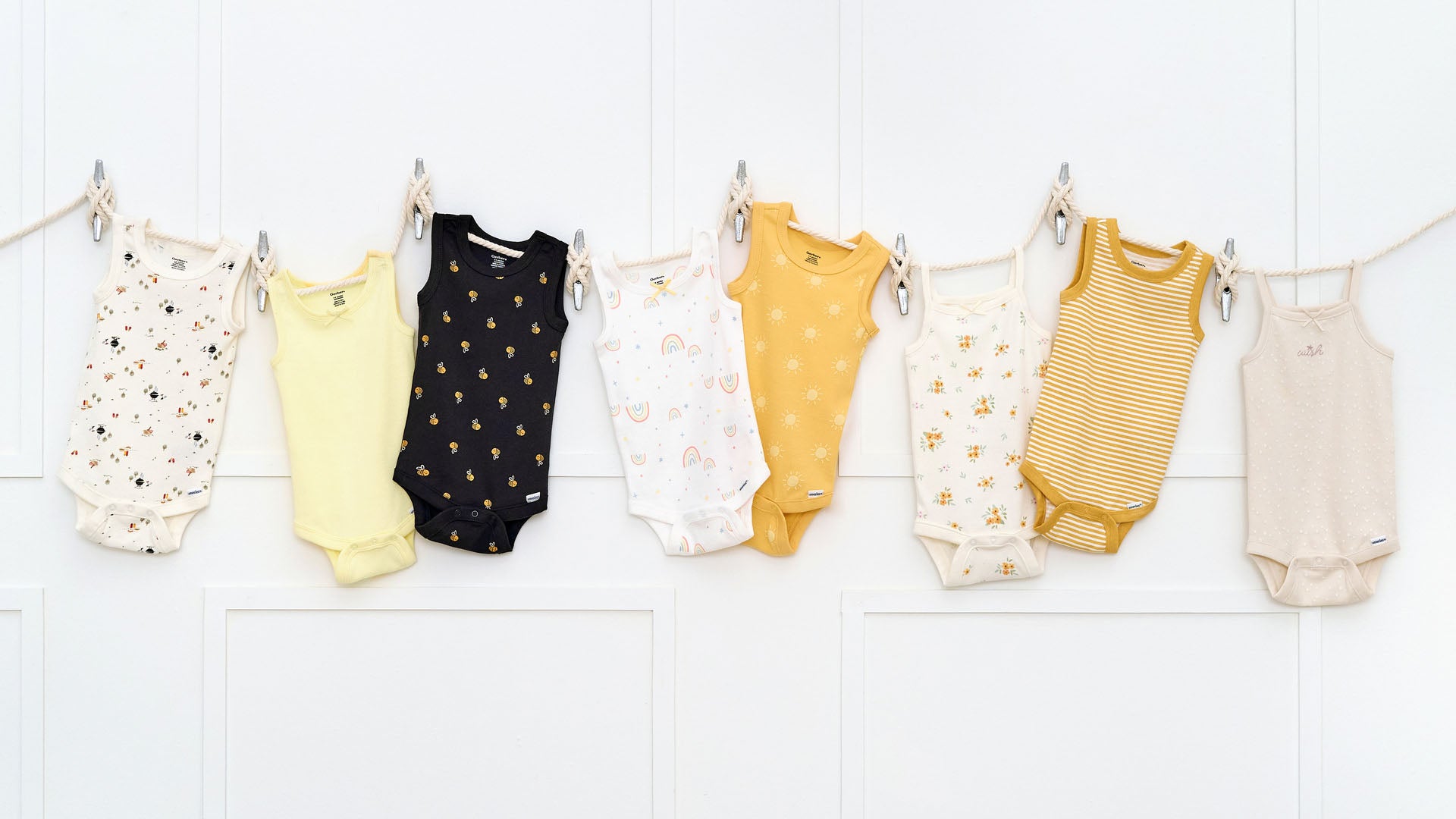 Seven baby bodysuits hanging in a row on a white wall, featuring various patterns and colors.