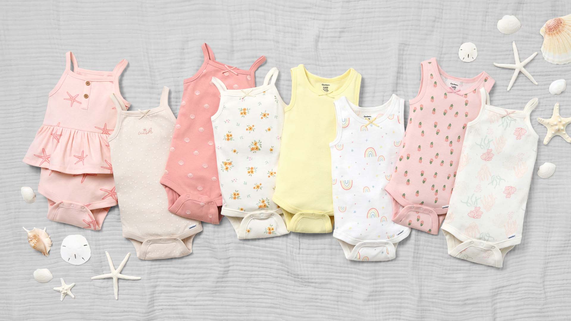 Eight baby onesies in various pastel colors and patterns laid out on a textured gray background, accompanied by small seashells and starfish.