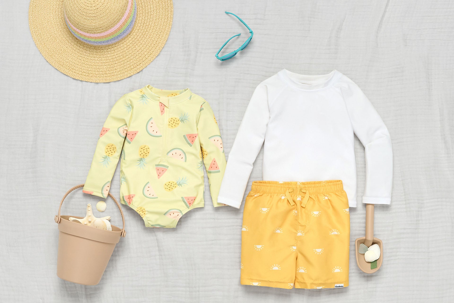 Flat lay of children's clothing including patterned girls swimsuit, boys white rashguard shirt, yellow swim shorts, with accessories like a straw hat, sunglasses, bag, and toy on a gray fabric background.