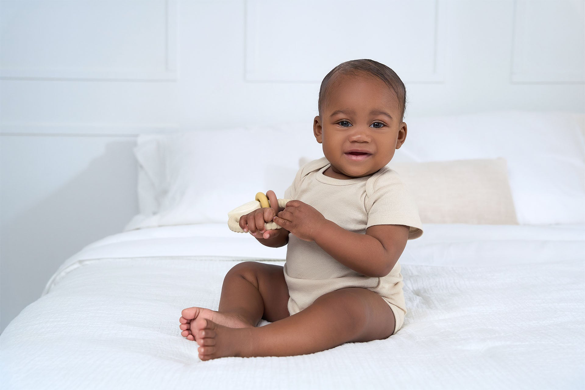 Baby sitting on a white bed, holding a toy, and smiling at the camera in a beige onesie bodysuit.