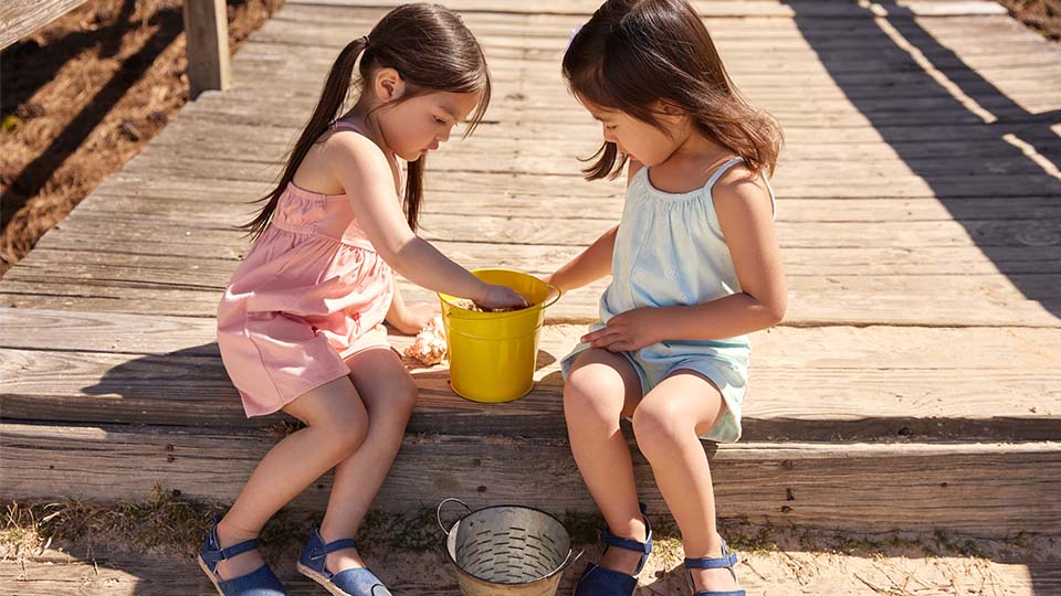 Two young girls sitting on a wooden boardwalk, playing with shells in a yellow bucket on a sunny day.