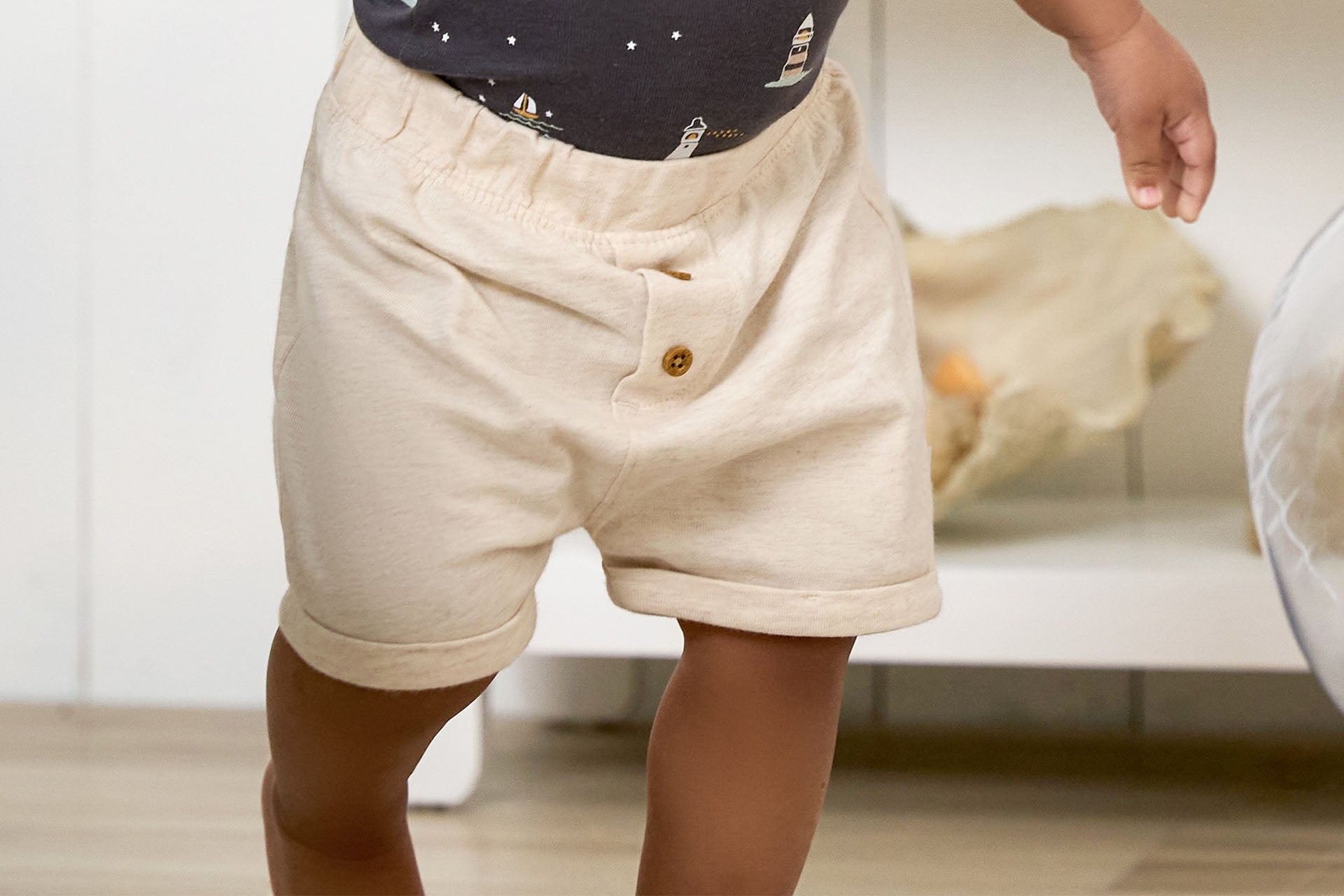 Close-up of a toddler standing in beige shorts and a patterned navy shirt, indoors, with only lower body and legs visible.