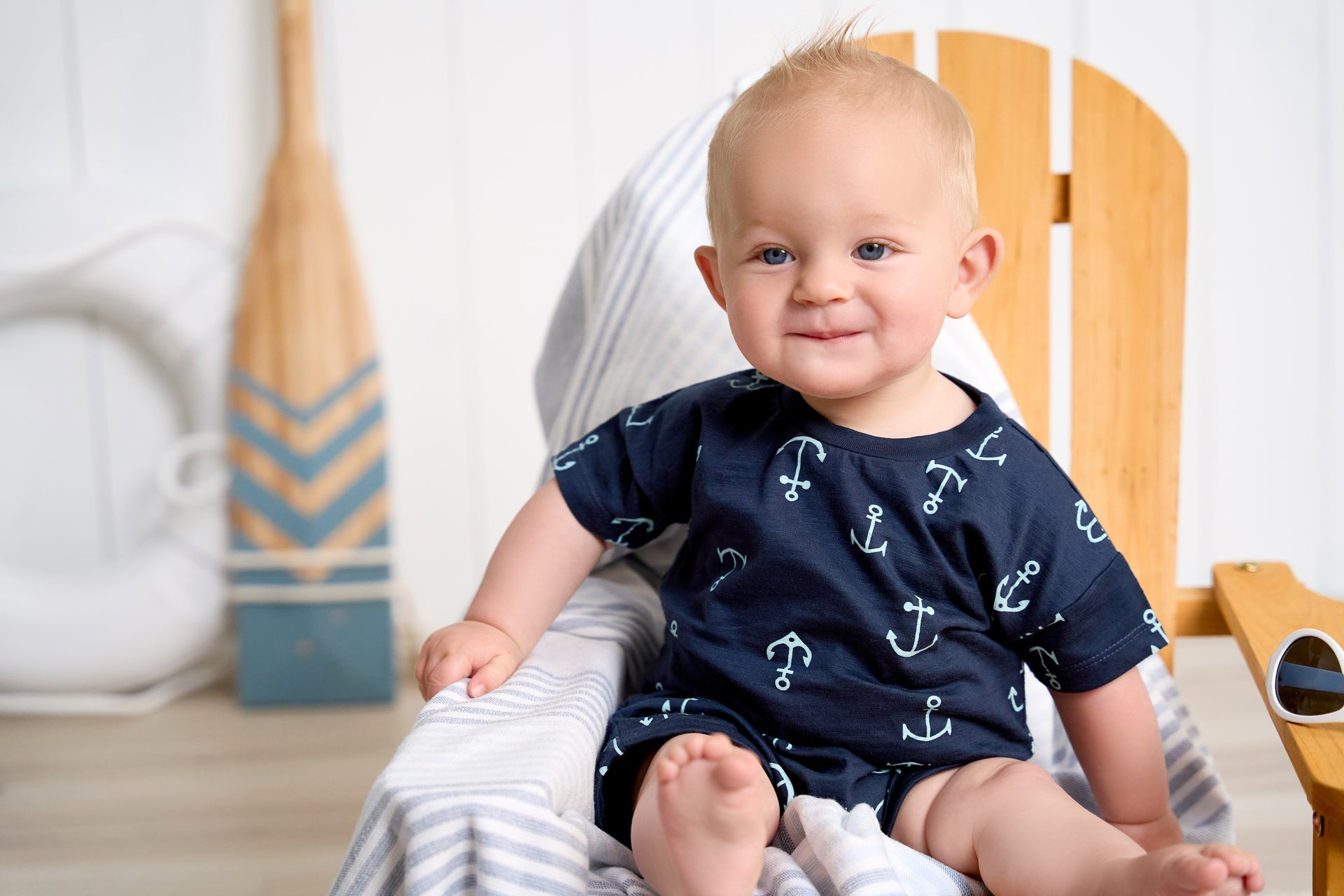 A baby boy sitting on a wooden chair, wearing a blue anchor-print shirt, smiling in a room with a striped cushion and a boat oar in the background.