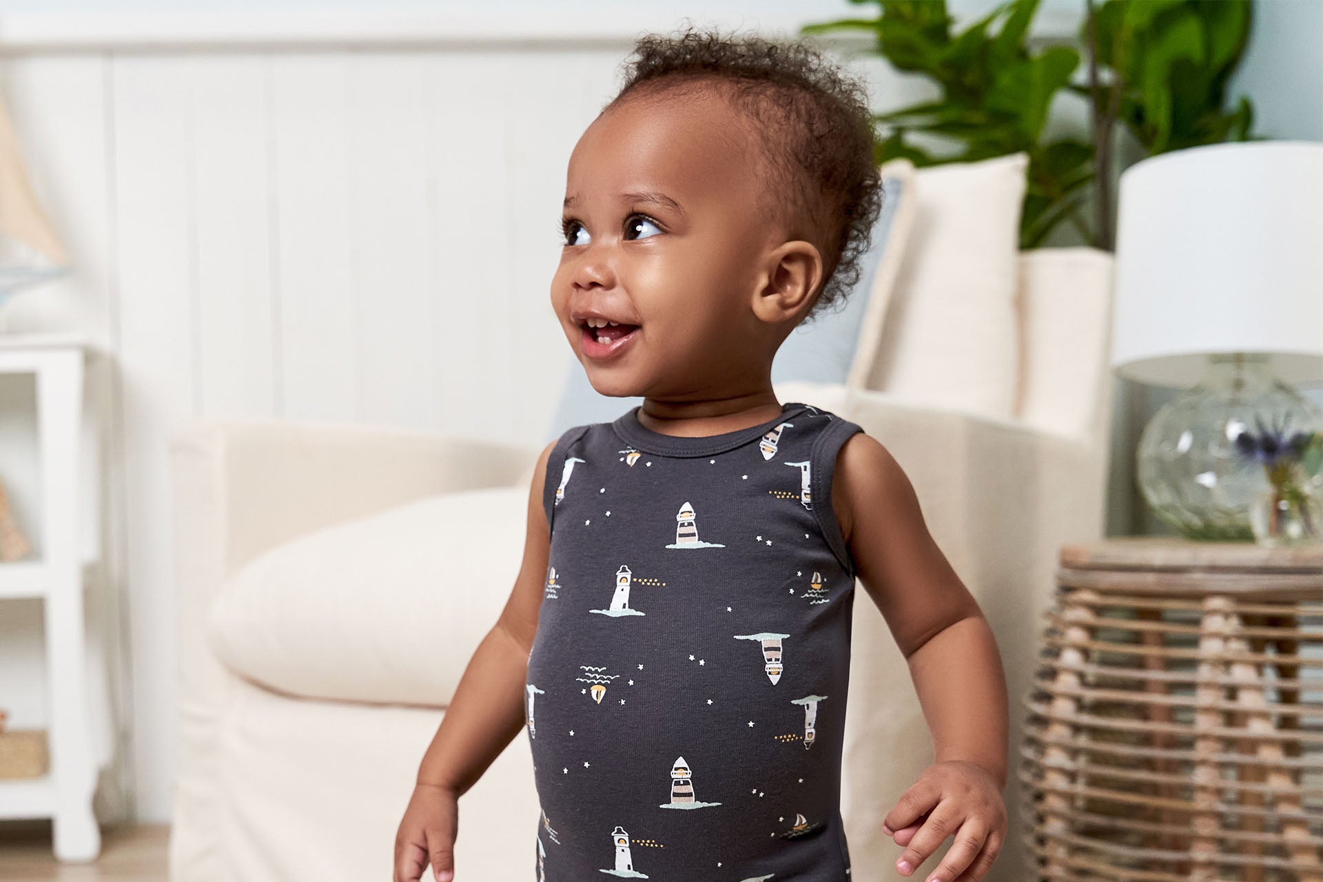 Toddler smiling in a room, wearing a dark onesie with lighthouse prints.