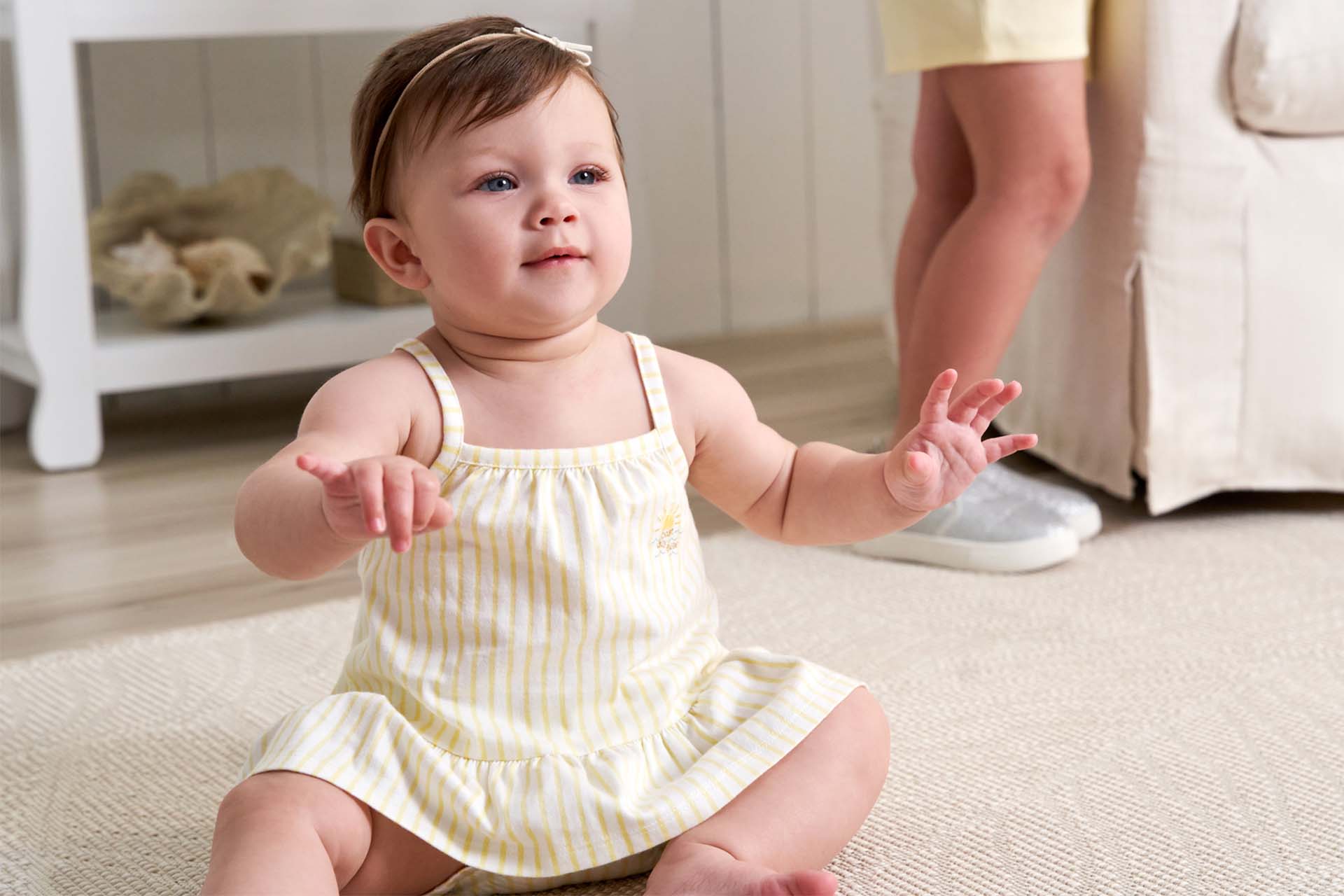 A baby in a yellow striped dress sits on the floor, looking up with arms outstretched, with a sibling standing nearby.