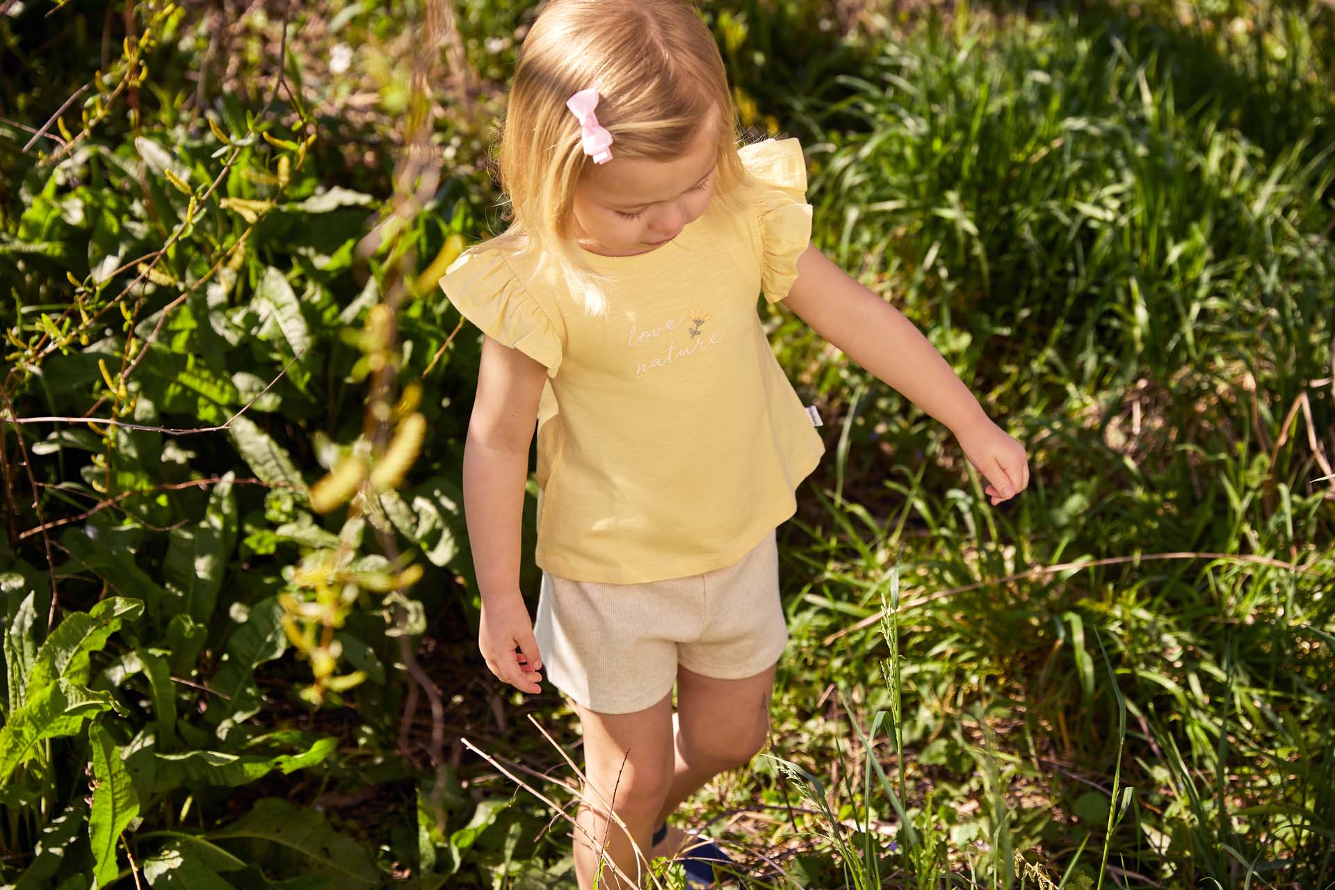 A young girl in a yellow shirt and beige shorts walking through a sunlit field with tall grasses.