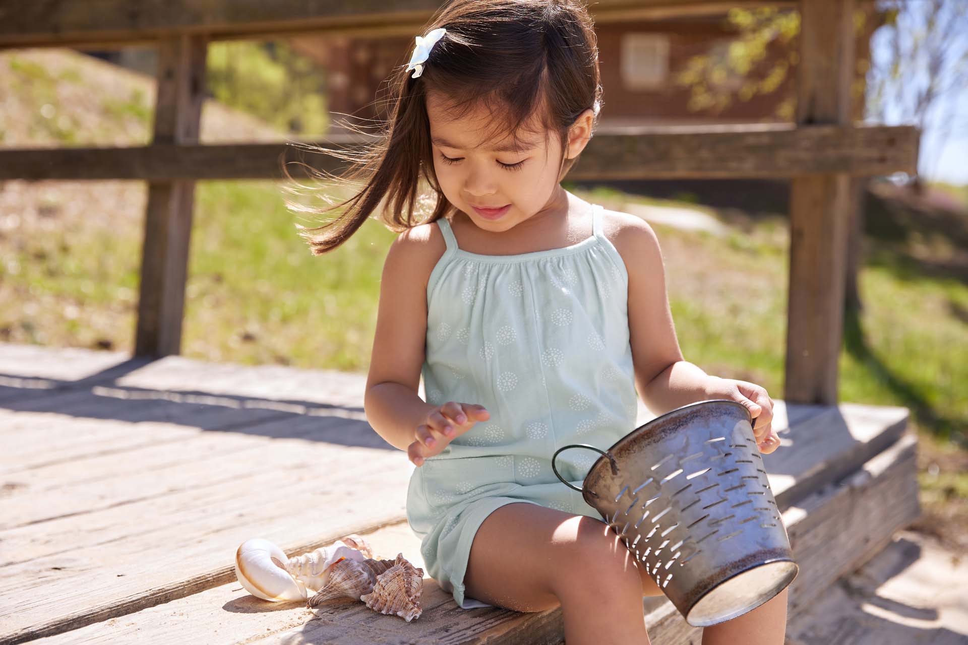 A young girl in a light blue romper sits on a wooden deck, examining seashells next to a metal bucket in a sunny, outdoor setting.