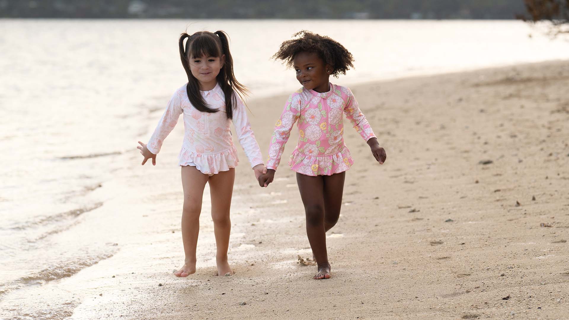 Two young girls in cute pink swimsuits holding hands and walking along a sandy beach in matching floral swimsuits.