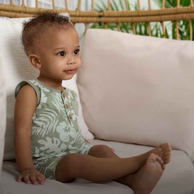 A baby in a green romper sitting on a wicker chair.