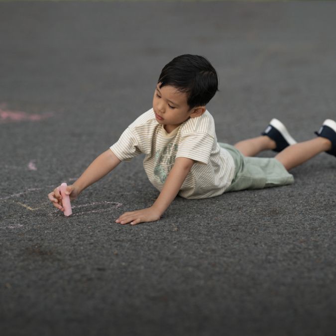A young boy laying on the ground and drawing with chalk.