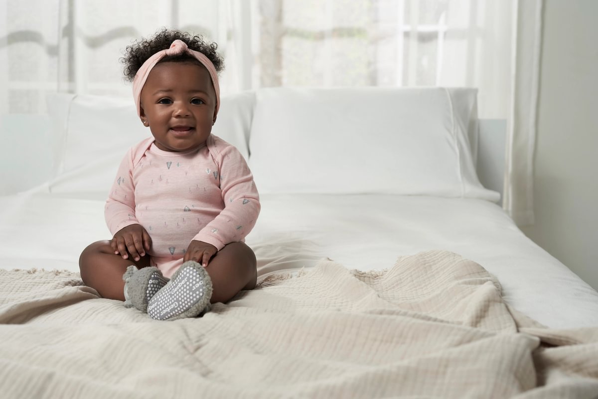 Baby sitting on a bed wearing a pink outfit with a headband.