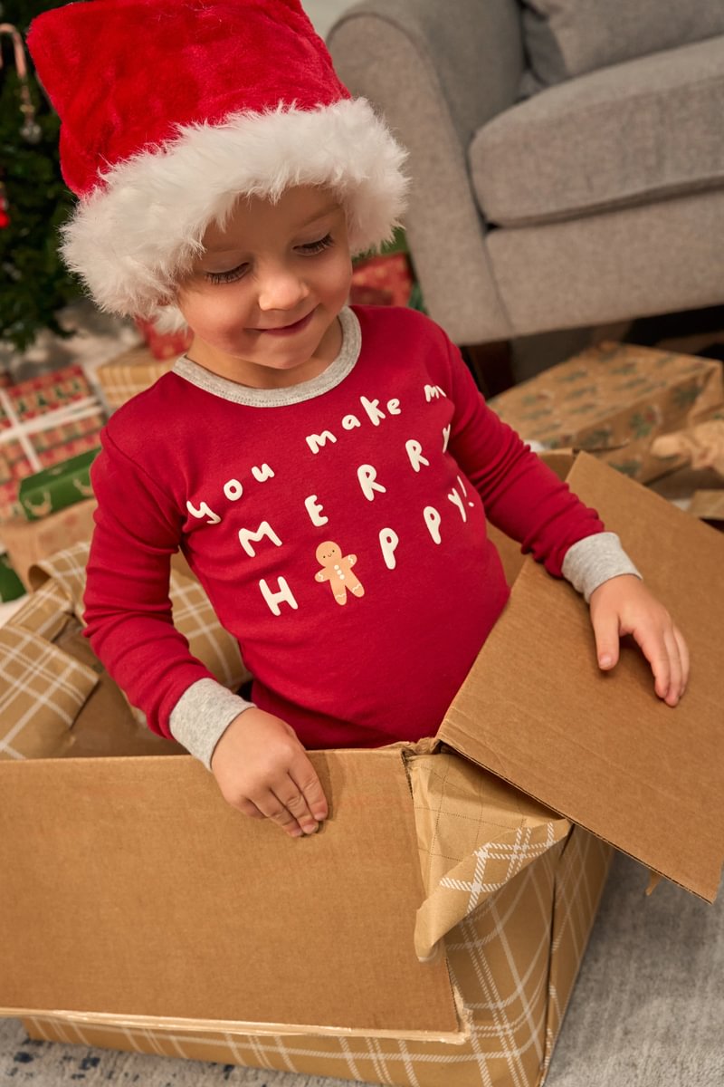 Child in a red Christmas sweater and Santa hat opening a cardboard box.