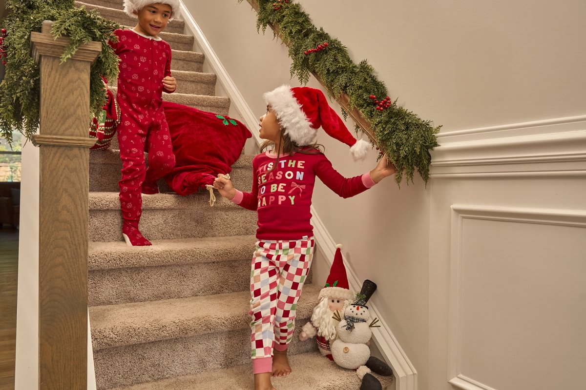 Two children in festive pajamas standing on a staircase with Christmas decorations.