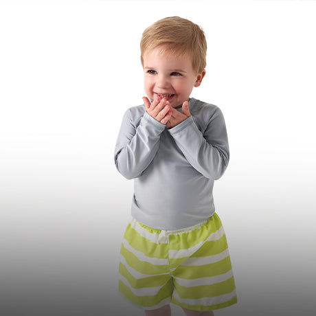 Boy giggling in yellow swim trunks laughing and holding hands, covering his mouth.