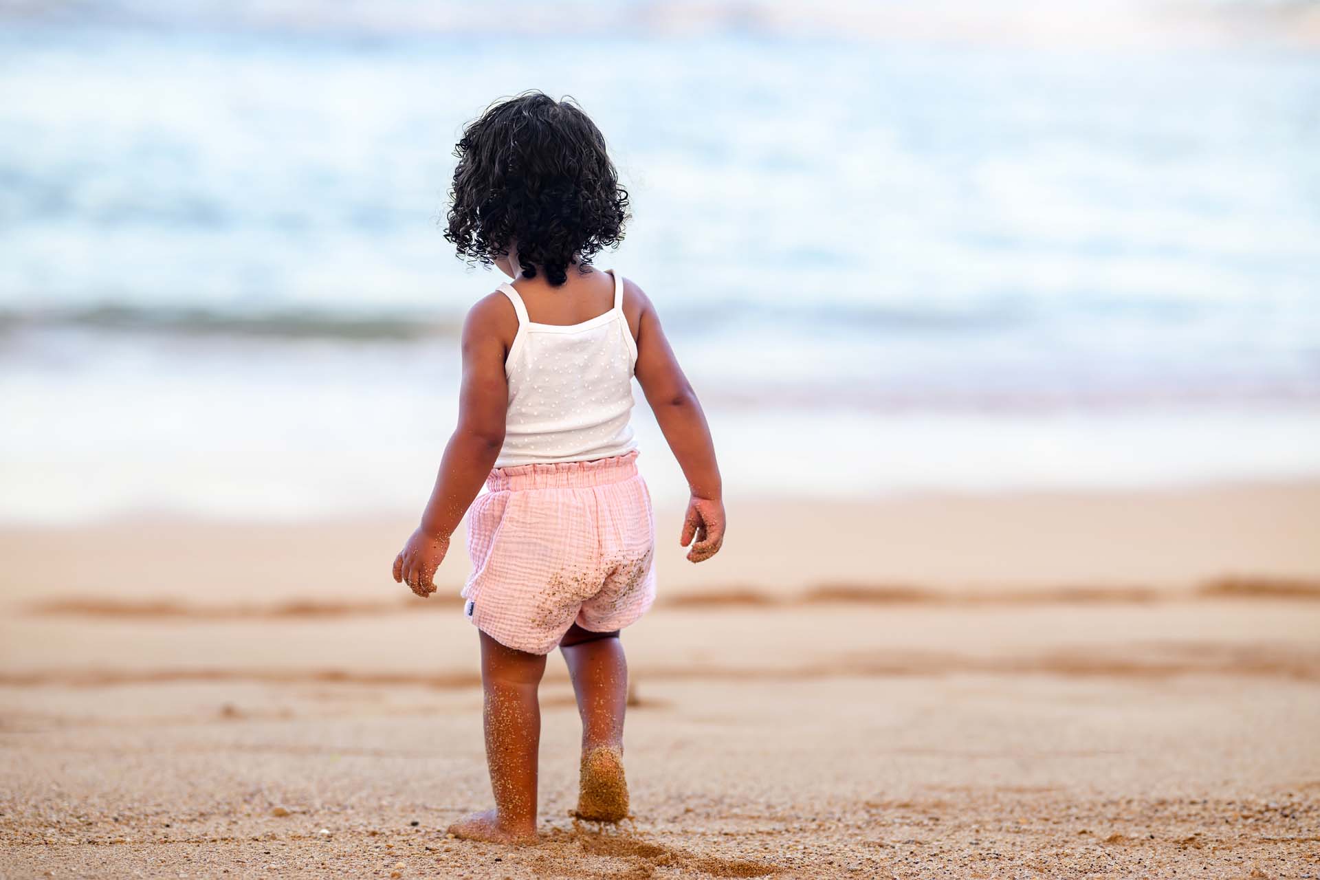 A young girl in a white tank top and pink shorts standing on a sandy beach, looking out at the ocean.