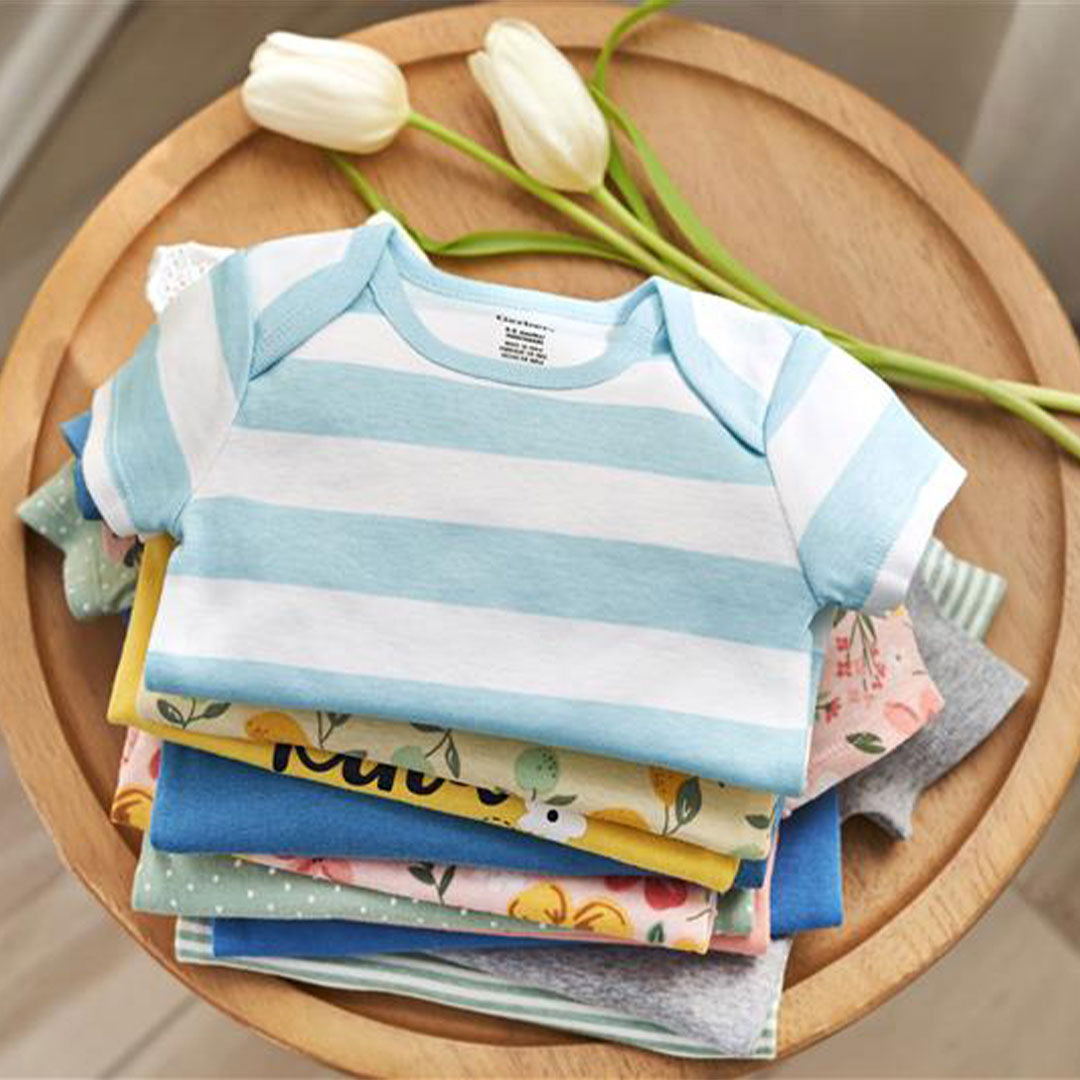A neatly folded stack of colorful baby onesies, including a striped blue and white piece, sitting on a wooden table adorned with fresh white tulips. Reflecting the fresh, cheerful spirit of our brand.