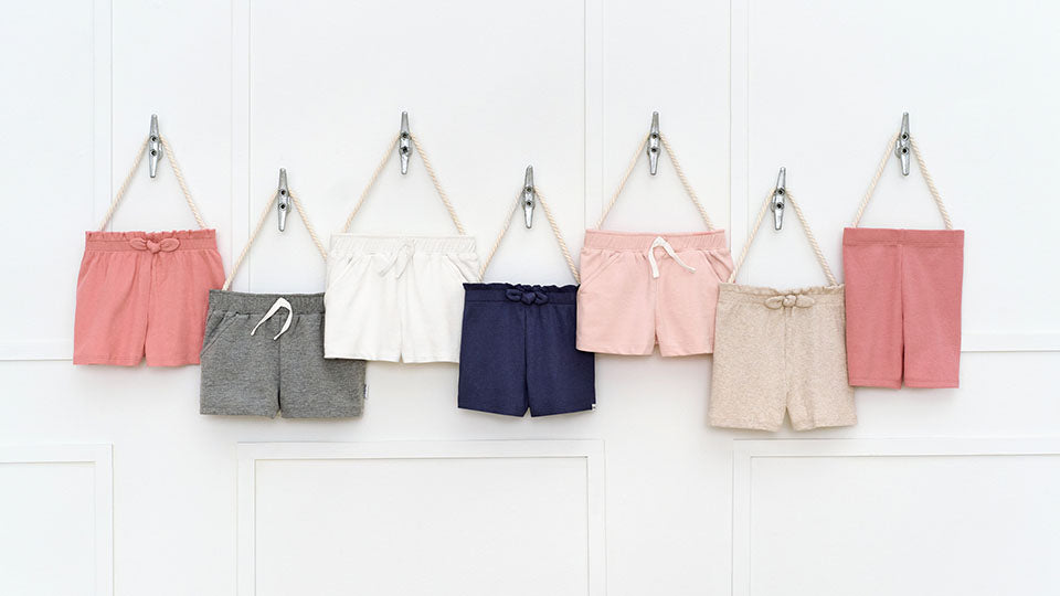 Seven pairs of toddler girl casual shorts in various summer colors hanging on hooks against a white wall.