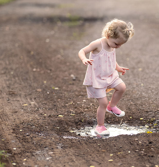 A young girl in a cute pink outfit & shoes splashing into a puddle on a dirt path.