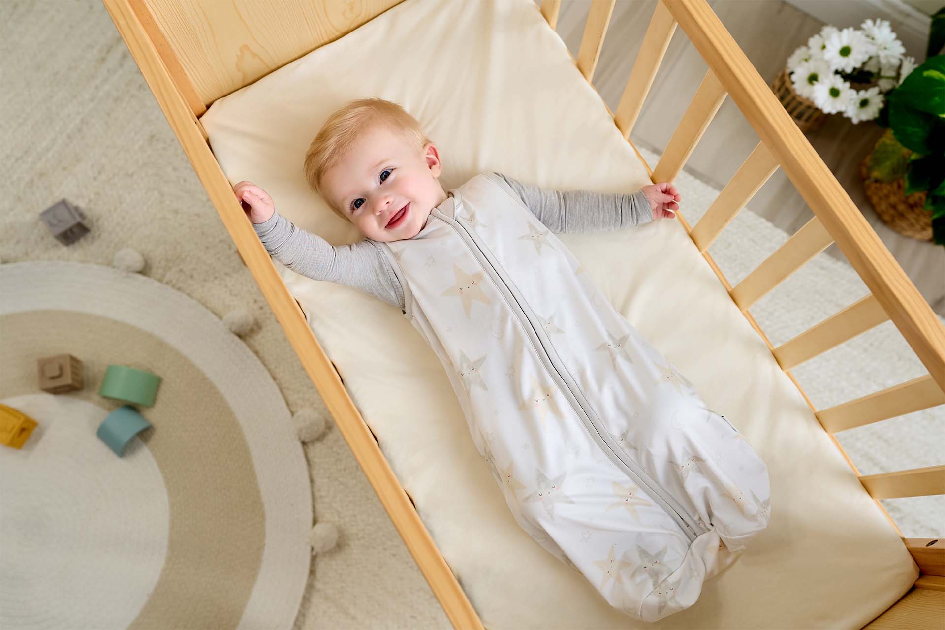 A baby in a Gerber Childrenswear sleep sack lying in a crib and smiling up at the camera.