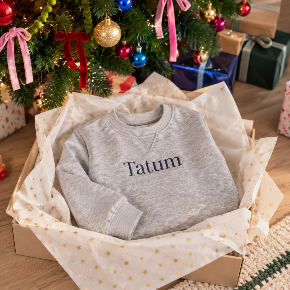 A light gray crewneck sweatshirt with the name 'Tatum' embroidered in navy blue across the chest, presented inside a gift box lined with tissue paper under a decorated Christmas tree.