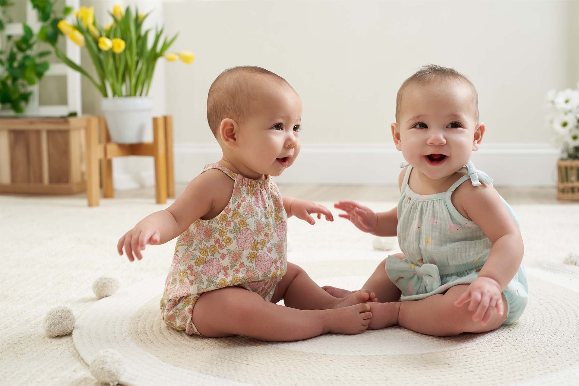 Two baby girls wearing spring outfits are sitting on a rug, smiling and playing with each other.