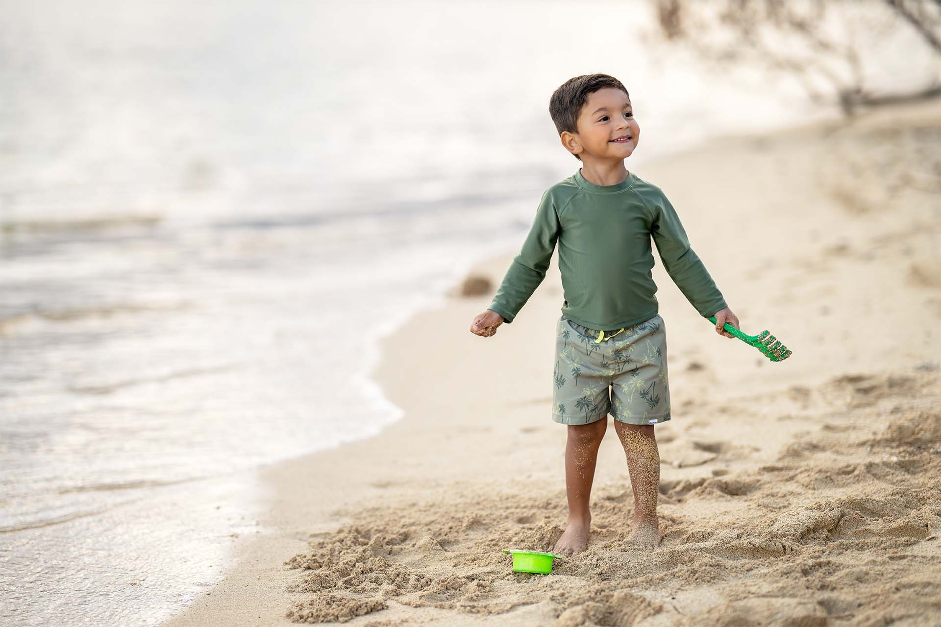 A toddler boy in a green rash guard shirt and green swim trunks with a toy shovel standing on a sandy beach looking cheerful.