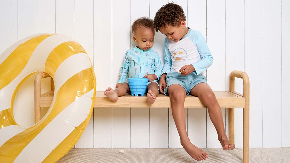 Two young boys sit wearing swimsuits on a wooden bench, playing with a blue bucket and toy, with an inflatable ring and white paneled wall in the background.
