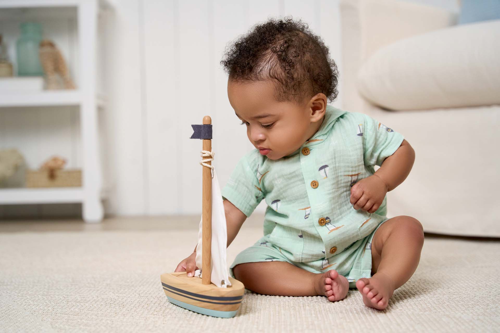A baby boy wearing a sailboat patterned romper examines a wooden toy boat in a living room.