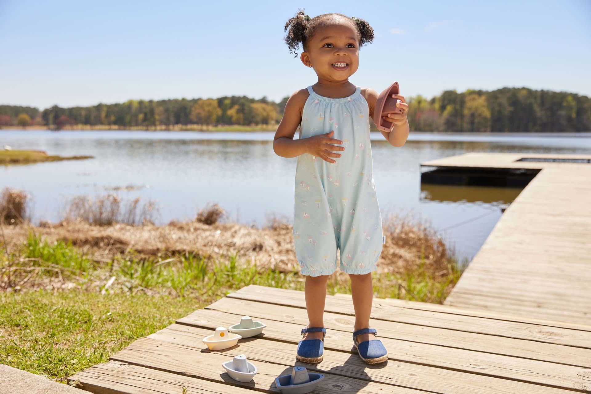 A toddler girl in a blue outfit smiles and points, standing on a wooden dock by a lake with toys nearby, on a sunny day.