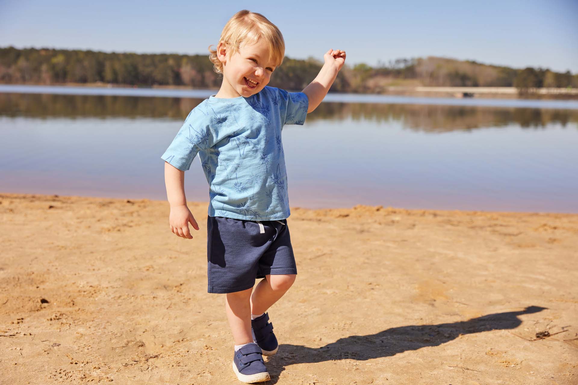 Young boy in a blue shirt and shorts standing on a sandy lakeshore, raising one arm in joy.