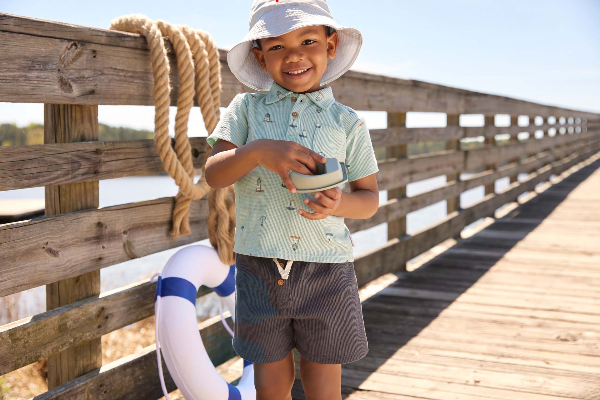 Young boy in a sun hat and summer outfit smiling and holding a toy boat on a wooden pier with a lifebuoy nearby.