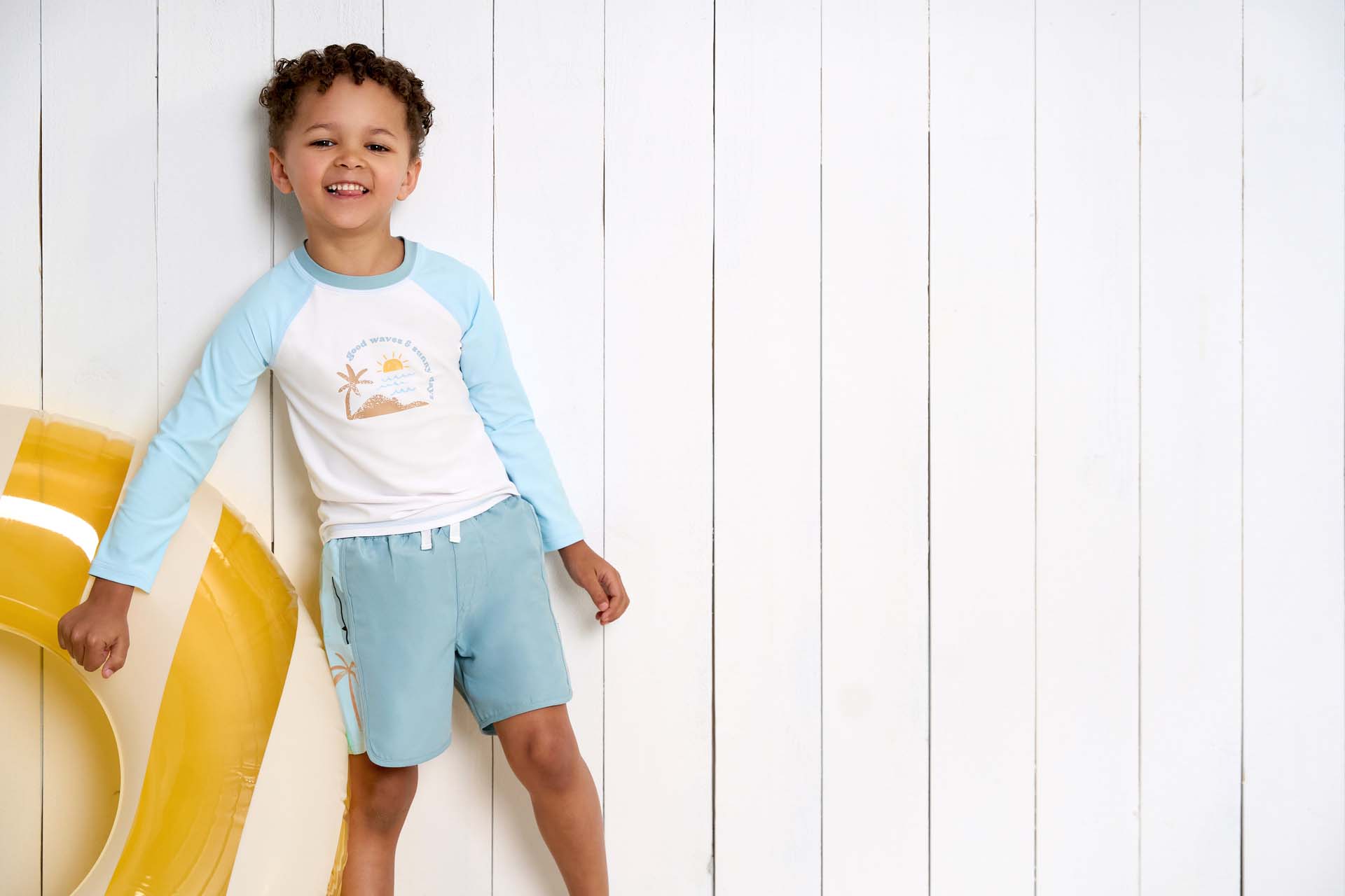 A toddler boy in a blue swim set smiling and posing with a yellow swim ring, standing against a white wooden background.
