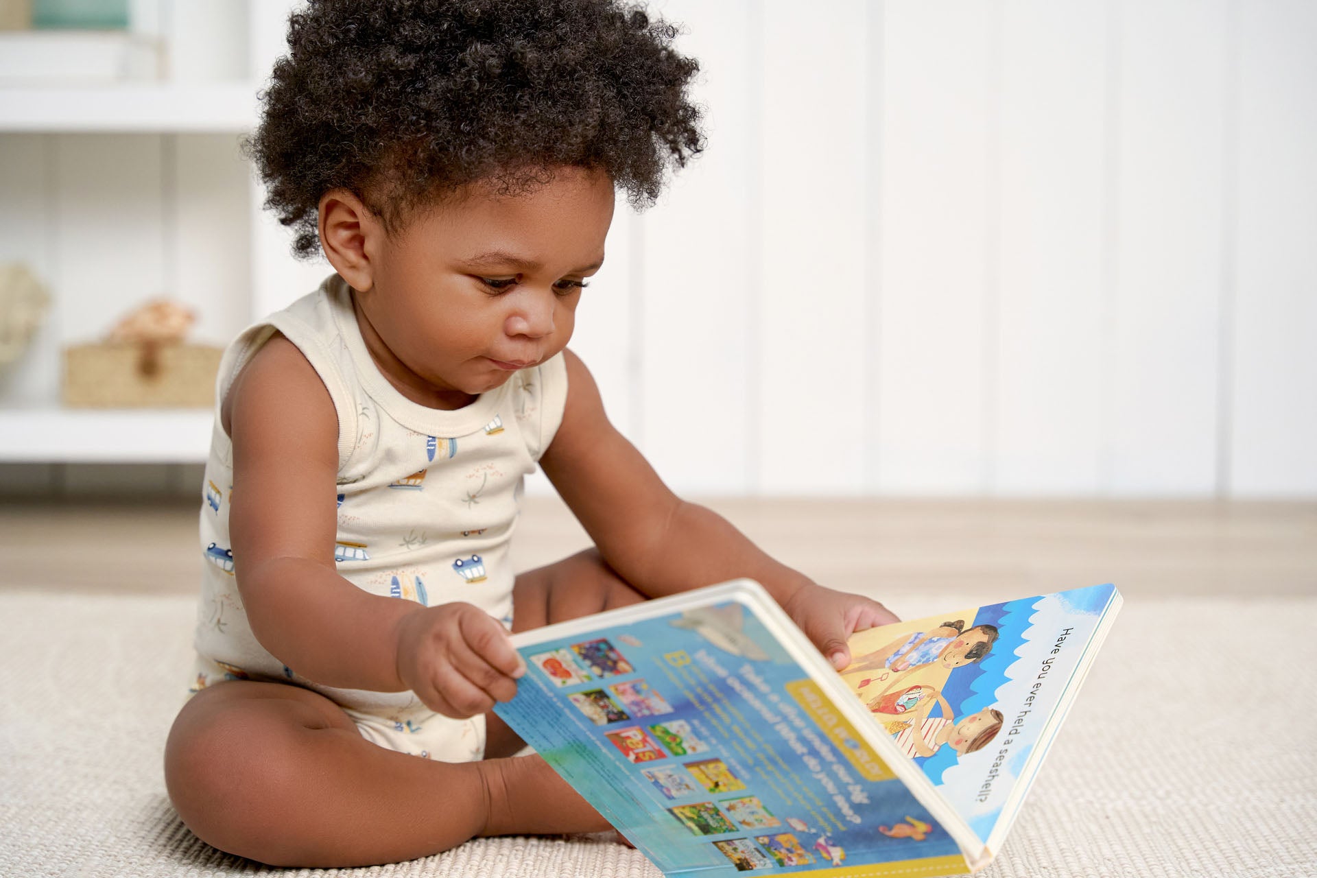 A toddler in a summer themed bodysuit sitting on a carpet, engrossed in reading a colorful picture book.