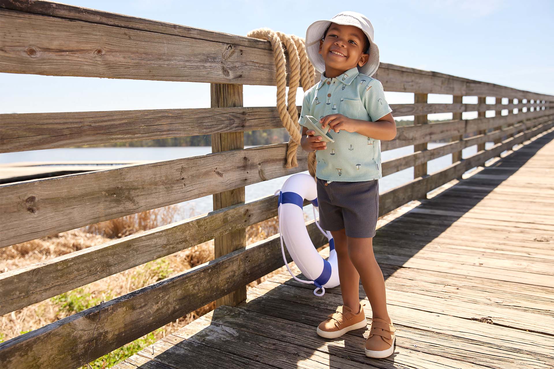 A toddler boy in a blue summer outfit standing on a wooden lakeside pier.