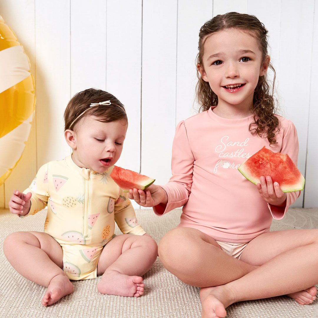 Two young girls in summer themed swimwear, the younger one in a headband and zippered swimsuit, the older in a pink top, both enjoying slices of watermelon.