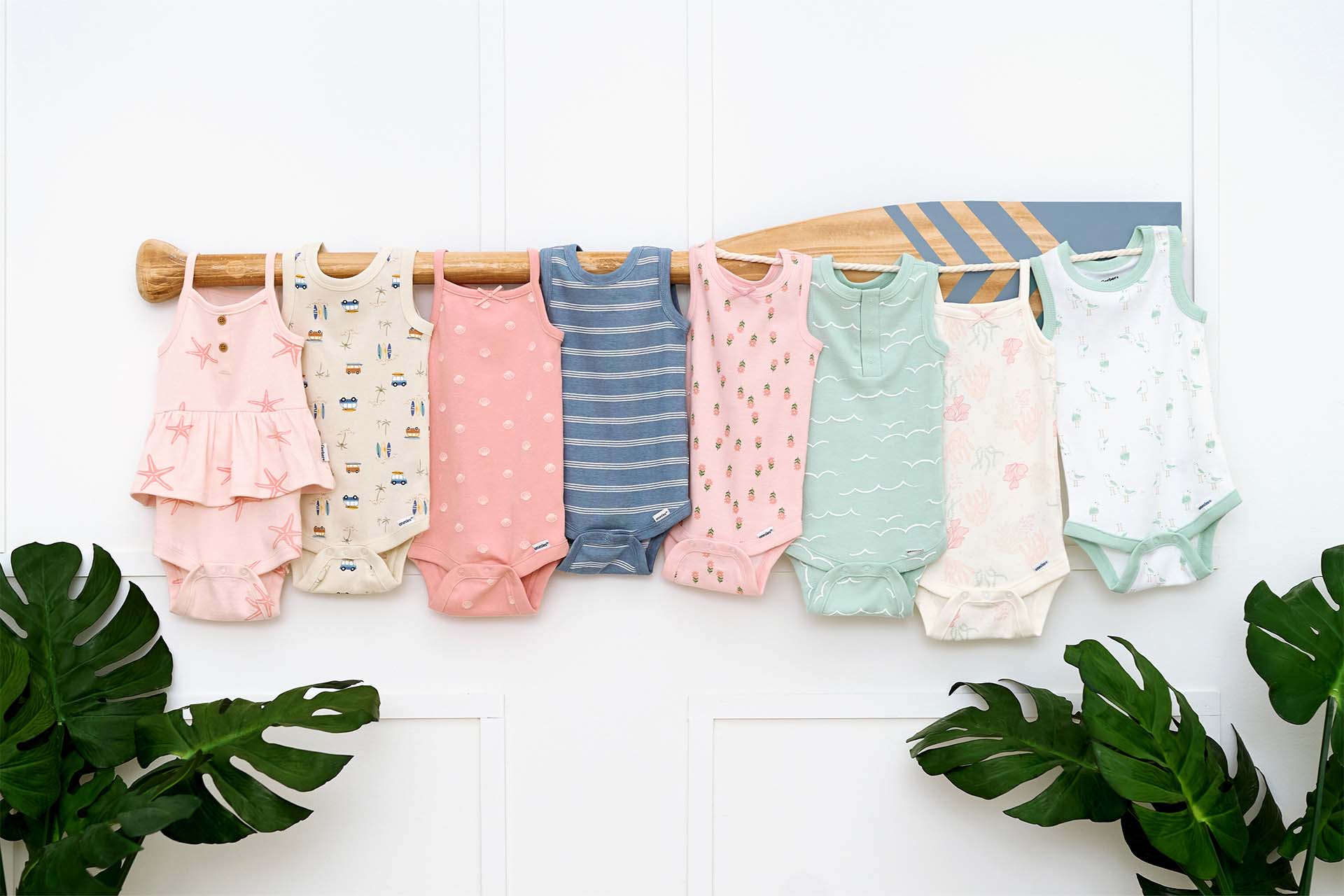 A row of baby onesies in various colors and patterns, including pink, white, blue, and green, hangs on a wooden rod. Two large green leaves can be seen in the bottom corners of the image.