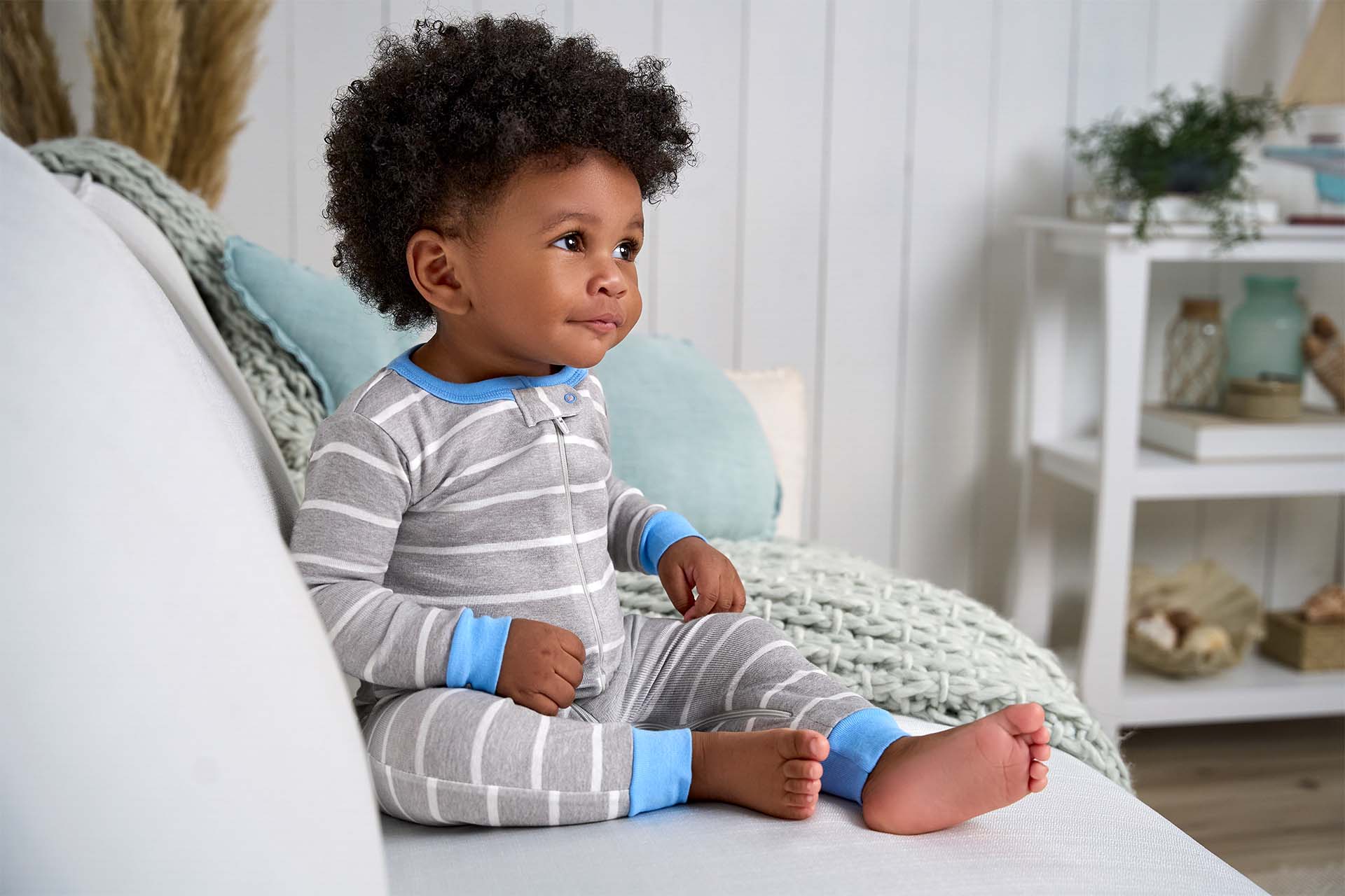 A baby with curly hair, dressed in a gray and white striped zip up pajama with blue cuffs, sits on a light-colored couch in a cozy, well-decorated room.