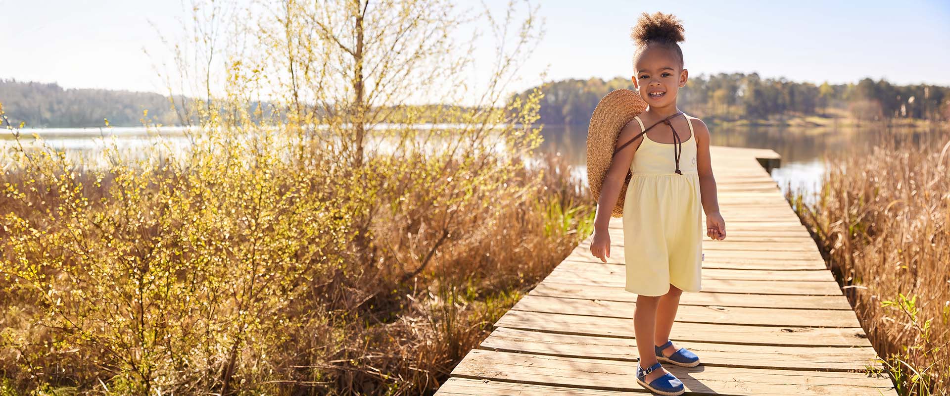 A young girl in a yellow romper and blue shoes stands on a wooden dock by a lake, holding a large straw hat. It is a sunny day with a clear blue sky with a lake in the background.