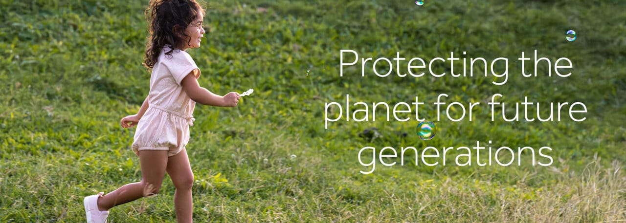 Smiling toddler girl running through green grass while blowing bubbles, wearing a light pink eco-friendly romper from Gerber Childrenswear’s sustainable baby clothing collection. Text overlay reads “Protecting the planet for future generations.” Shop Gerber Childrenswear for affordable sustainable baby clothes, toddler outfits, and eco-conscious fashion deals.