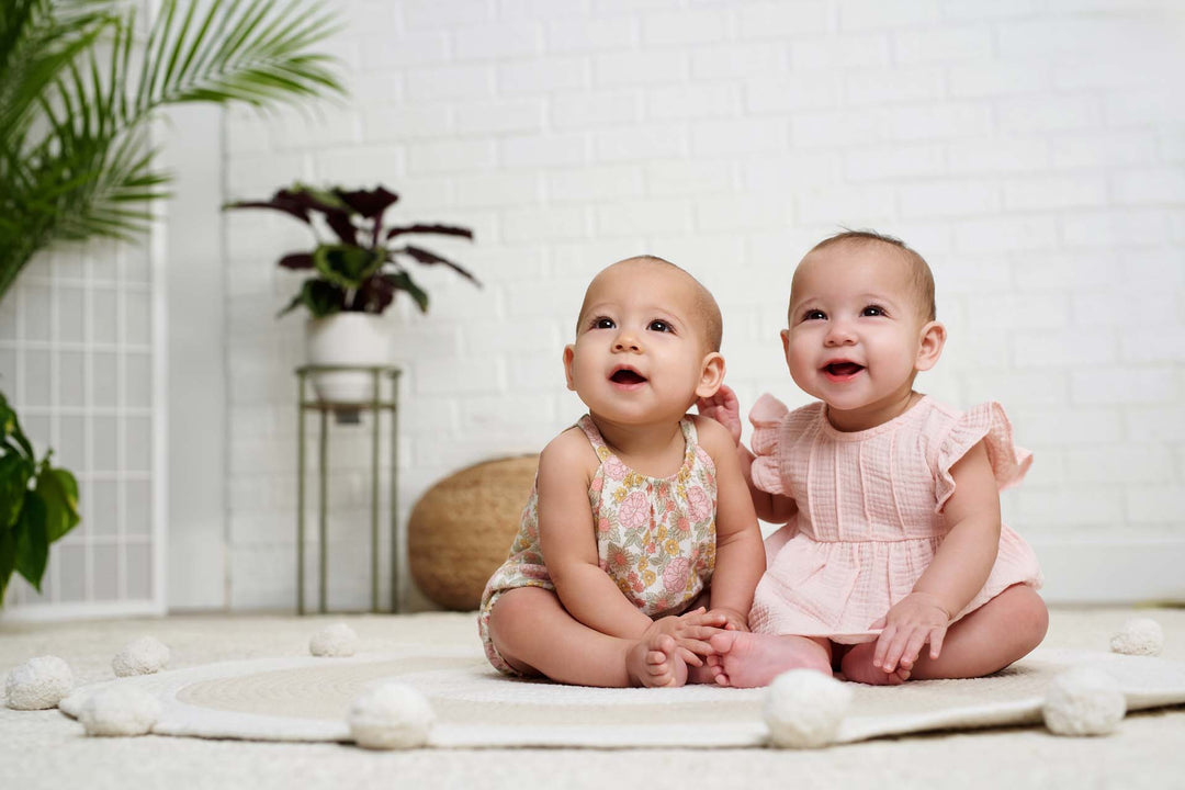 Twin baby girls. The left one has on a floral outfit and the right one has on a pink dress.