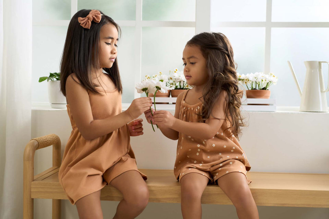Two little girls wearing coordinating outfits sitting on a bench and holding flowers.