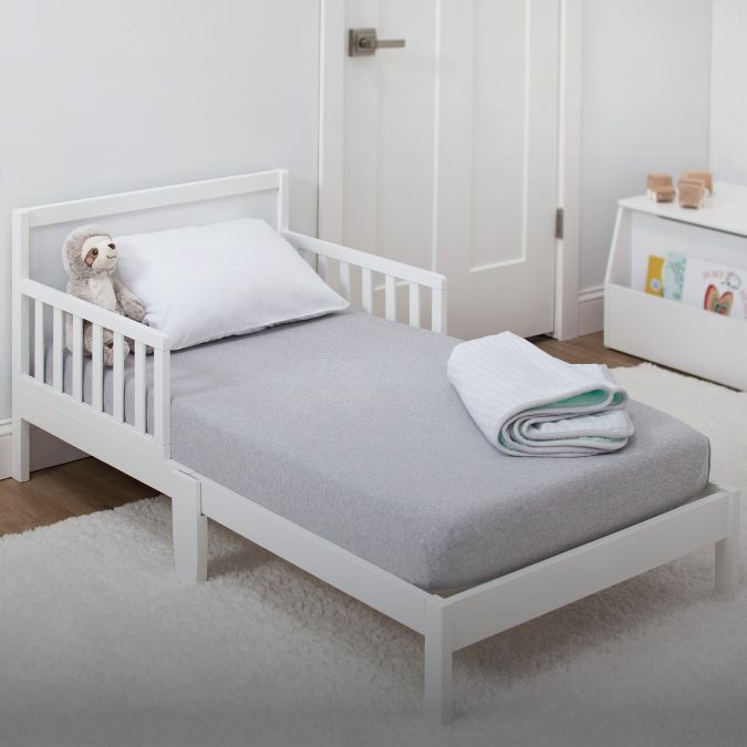 A white toddler bed with grey bedding in a room with a teddy bear.