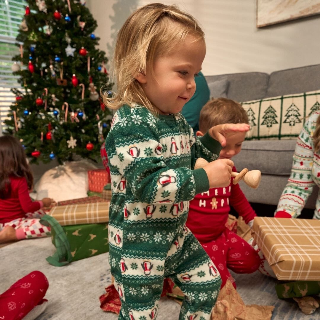 Children in festive pajamas playing on a carpeted floor with a decorated Christmas tree in the background.