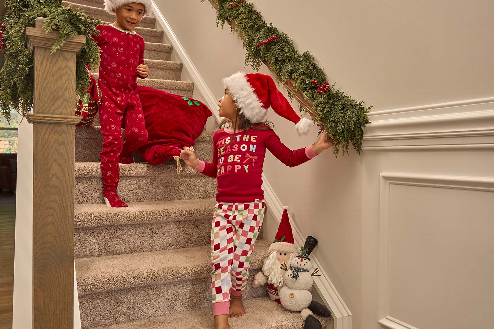 Two children in festive pajamas on a staircase with Christmas decorations.