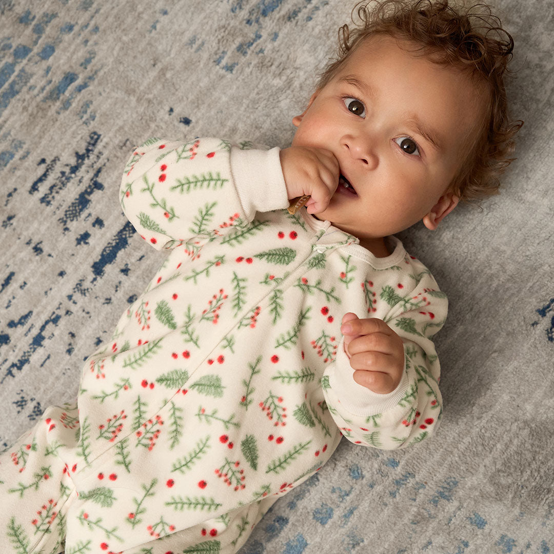 Baby wearing a white onesie with green leaf and red berry pattern, lying on a textured blue and gray surface.