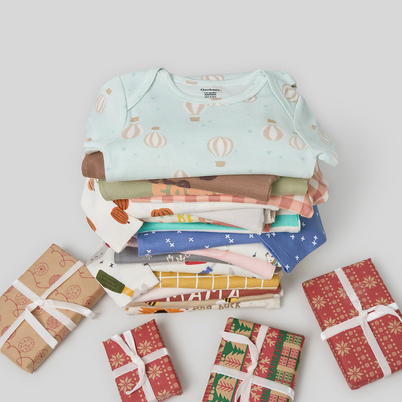 Stack of patterned baby clothes with small gift boxes on a light background