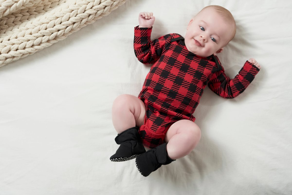 Baby wearing a red and black checkered outfit with black socks on a white surface.