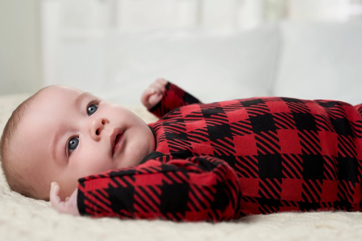Baby wearing a red and black checkered outfit lying on a white surface.